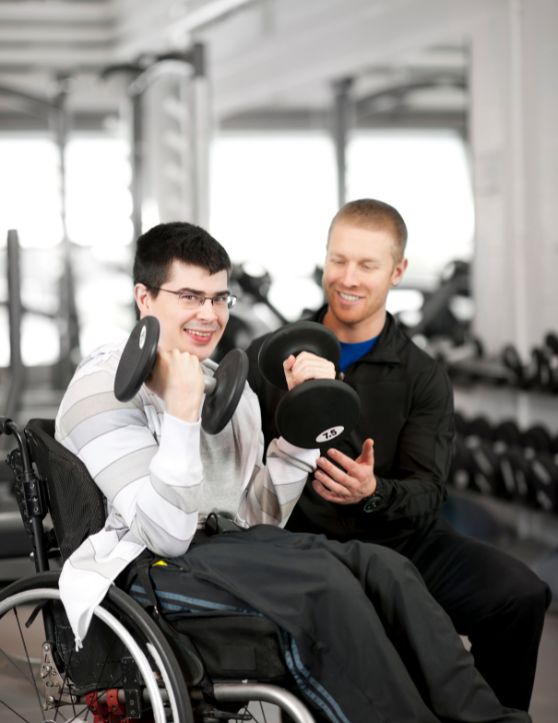 a young man in a wheelchair is lifting dumbbells with the assistance of a personal trainer in a gym setting