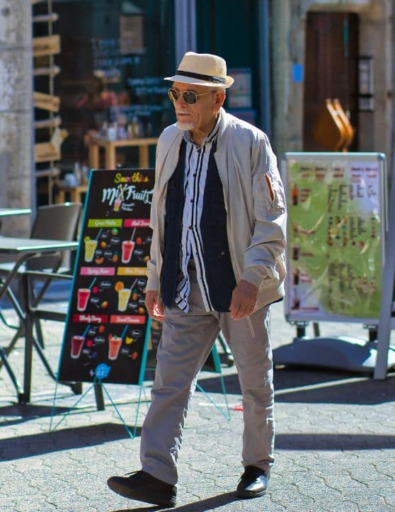 An elderly man, dressed in a white and black shirt with grey trousers, strolls along the sidewalk situated between two buildings. He accessorizes his outfit with a stylish hat and sunglasses. 