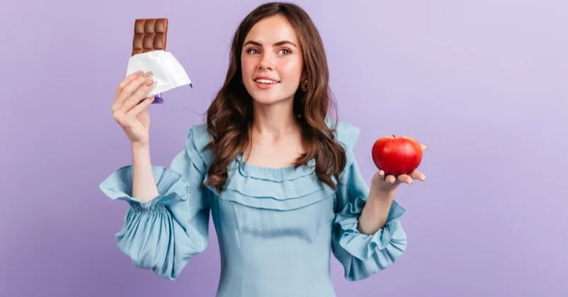 A beautiful green-eyed curly brunette girl is striking a pose with red apple and bar of milk chocolate in purple wall background