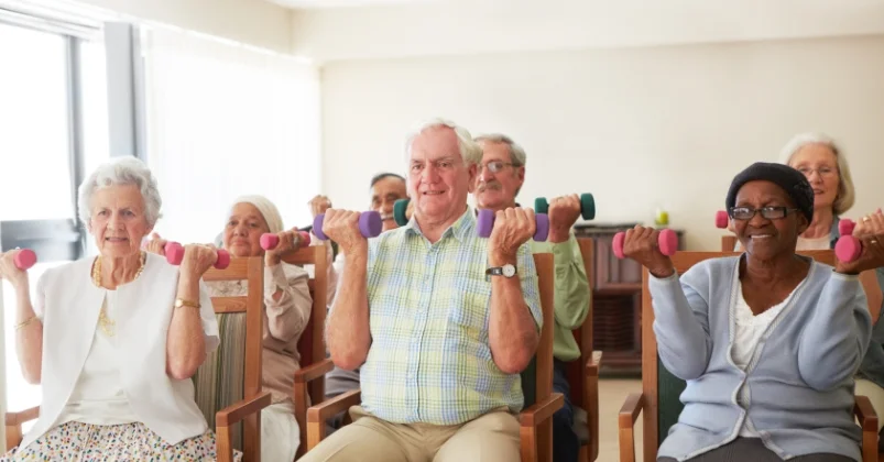 A group of senior individuals are engaged in seated exercises, utilizing dumbbells, within a room