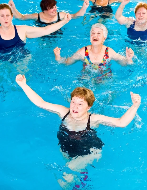 A group of senior women is engaging in water aerobics, raising their hands up in synchronized movements