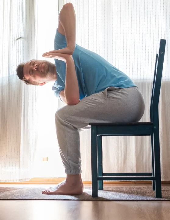 A man wearing a blue t-shirt sits on a chair, practicing yoga with his hands clasped behind his head, eyes closed