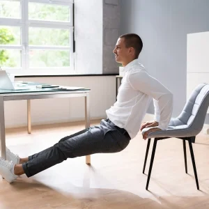 A man wearing a white shirt and grey pants is performing a Chair Plank exercise in a room 1