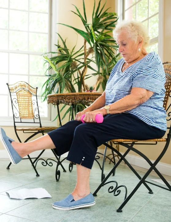 An elderly woman in a white shirt and black pants sits in a chair with hands on lap, holding a pink dumbbell, feet grounded in brown shoes