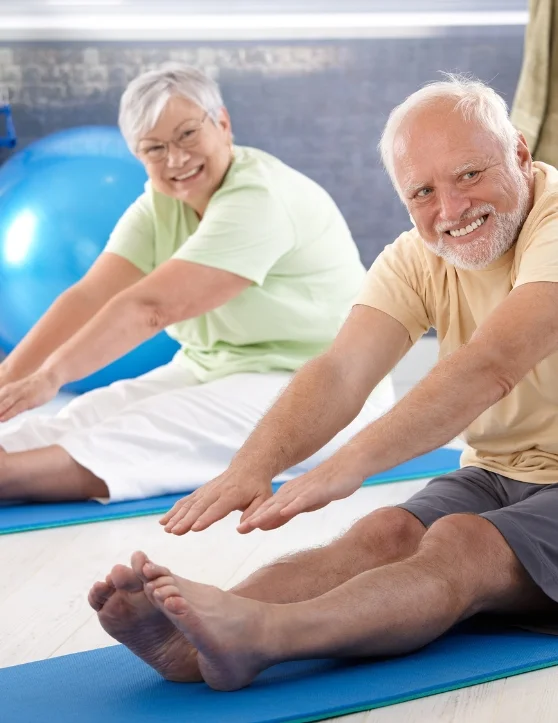 The elderly individuals are engaged in hamstring stretch exercises while seated on a blue mat adorned with smiley faces