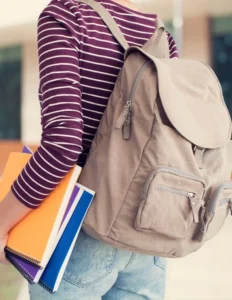 The girl is wearing a striped t-shirt, carrying a backpack on her back. She has books in her hands, held by her sides