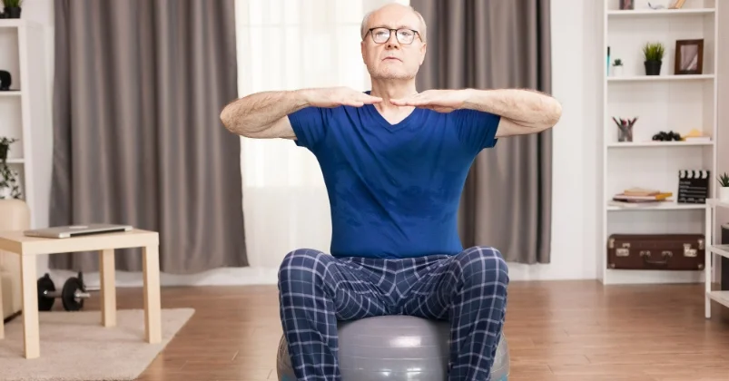 The senior man dressed in a blue t shirt and checked pants is exercising with a stability ball in his living room