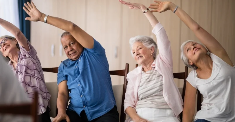 A group of elderly individuals are participating in chair exercises side bending as they raise their hands above their heads