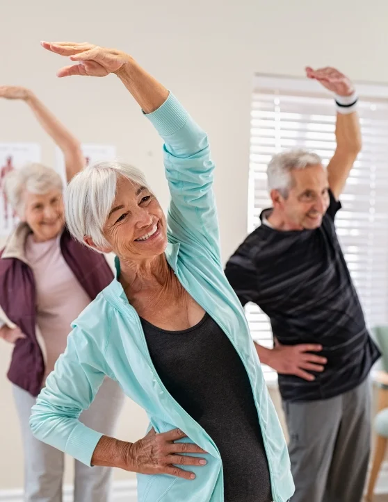 In a room, a group of elderly individuals are joyfully engaging in exercises, their smiling faces radiating happiness as they raise their hands above their heads to one side in unison