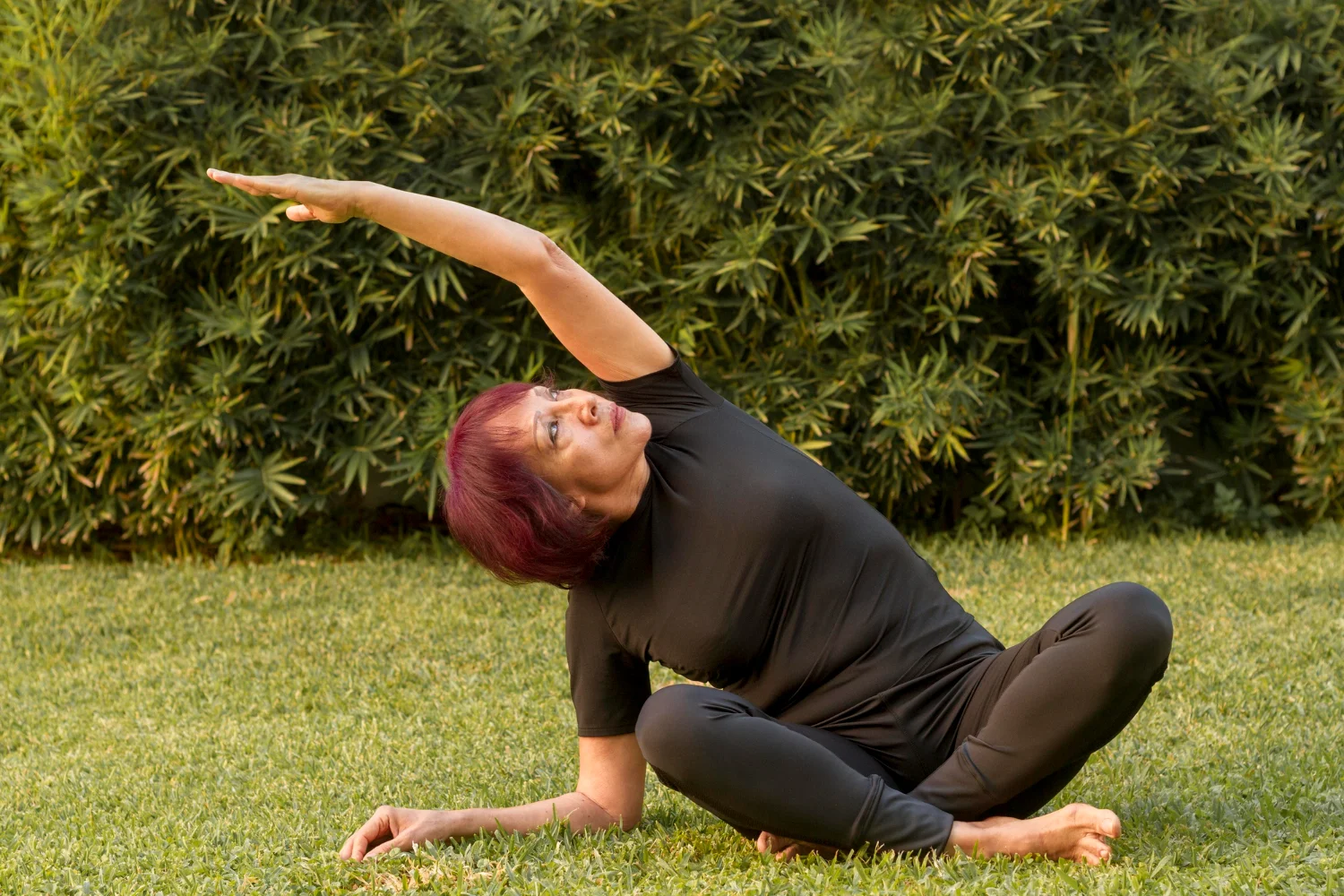 The elderly woman is seated on the grass dressed in a black outfit as she extends her hands above her head gently stretching to one side while practicing yoga