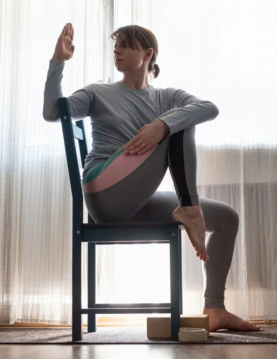 The woman is performing seated chair exercises, with one leg placed on the chair and the other on the floor. She turns her head towards the back while raising her hand and placing it on the top of the chair