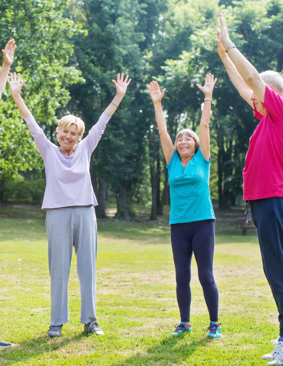 A group of elderly individuals is engaged in laughter yoga warm-up exercises in a natural environment