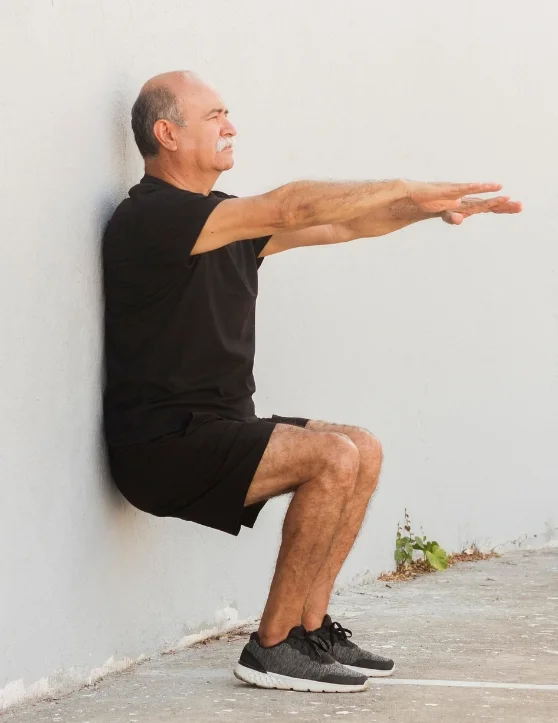 An elderly man dressed in black attire is engaged in a wall stand fitness exercise. He has his hands straight and is lightly leaning back against the wall, maintaining a seated position