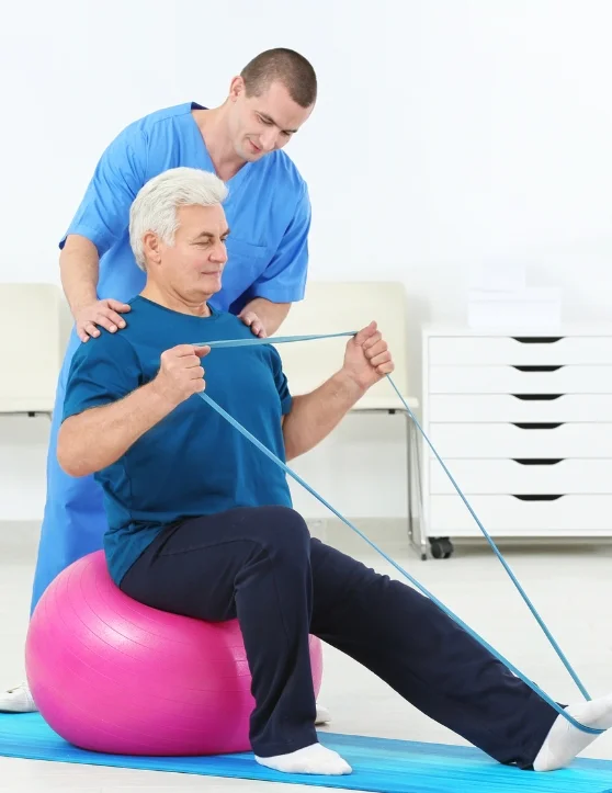 The elderly man is engaging in Swiss ball exercises under the guidance of a physical trainer