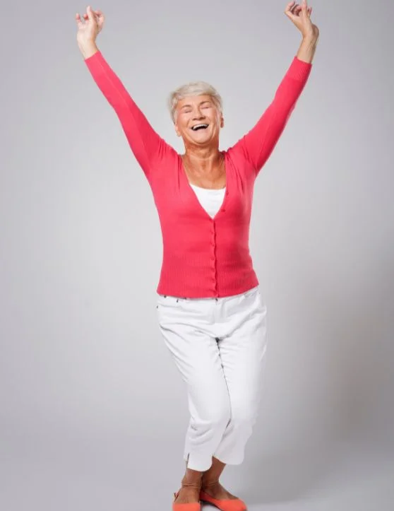 The elderly woman, adorned in a vibrant red top and white pants, joyfully raises her hands while swaying her body