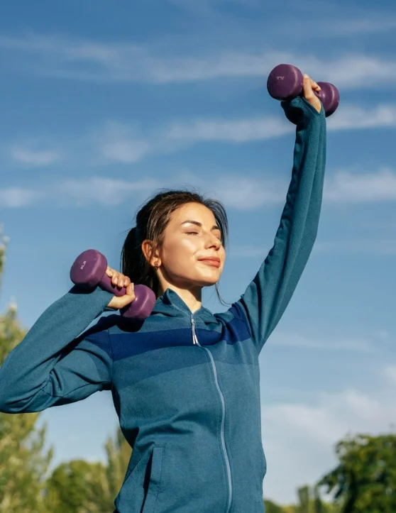 The girl, dressed in a gym suit, is exercising outdoors with dumbbells, her eyes closed, against a backdrop of the sky