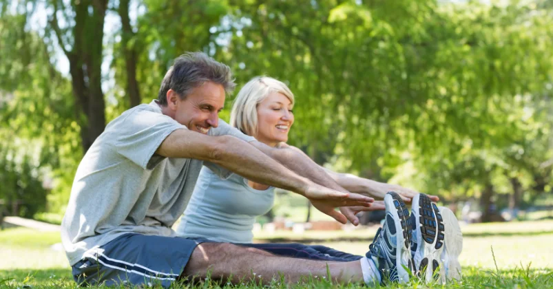 Two people are doing outdoor exercises, sitting on the grass, and attempting to touch their feet with smiling faces