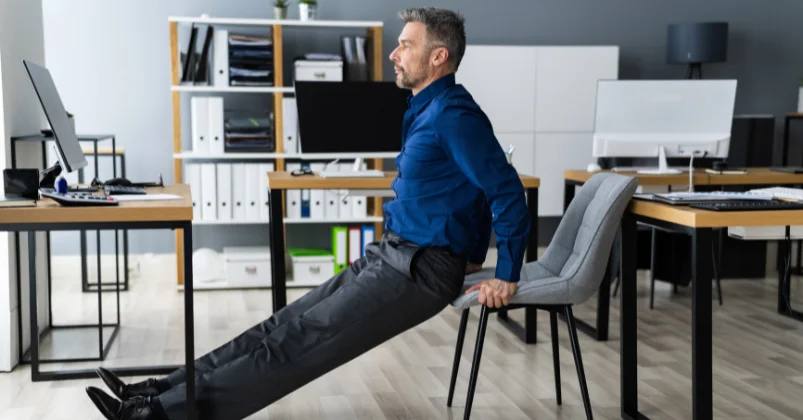 A man dressed in a blue shirt and dark grey pants is performing chair exercises in an office setting. He lands on his legs with impeccable form keeping them straight as he touches the floor