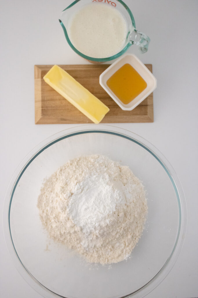 all-purpose flour and baking powder in a glass bowl along side a stick of frozen butter, honey and buttermilk