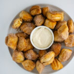 cinnamon sugar coated biscuit bites placed on a glass plate along with white sauce placed in middle