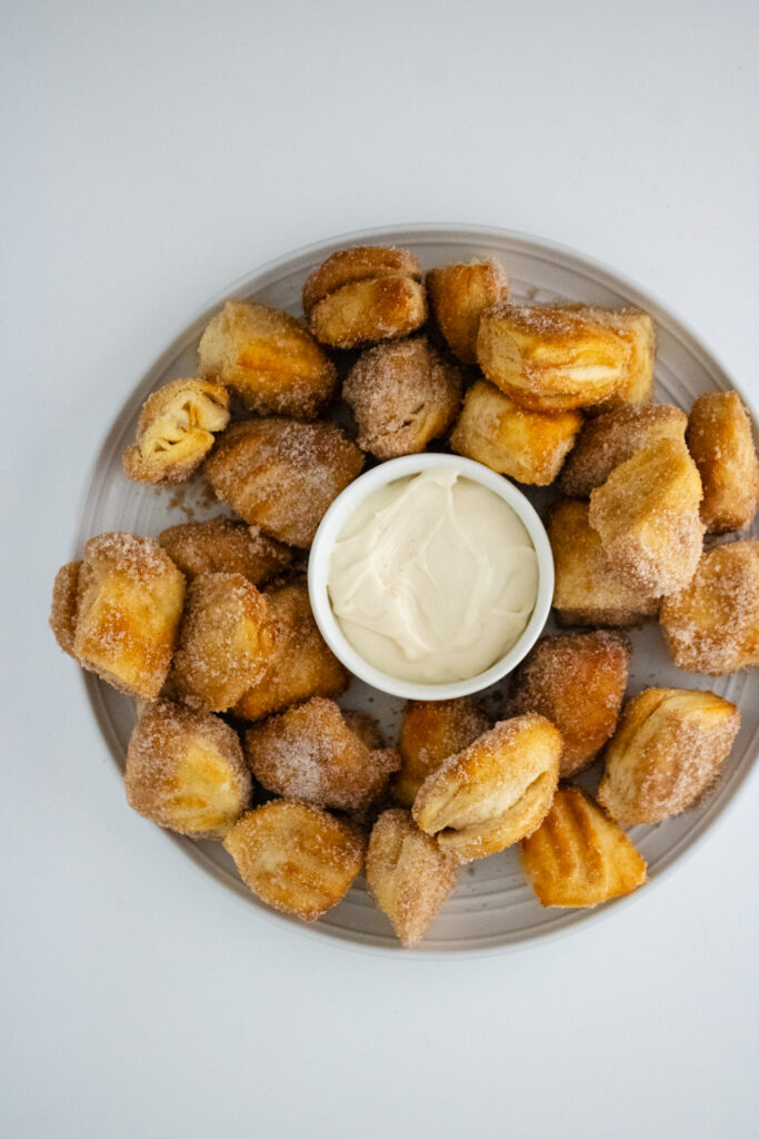 cinnamon sugar coated biscuit bites placed on a glass plate along with white sauce placed in middle