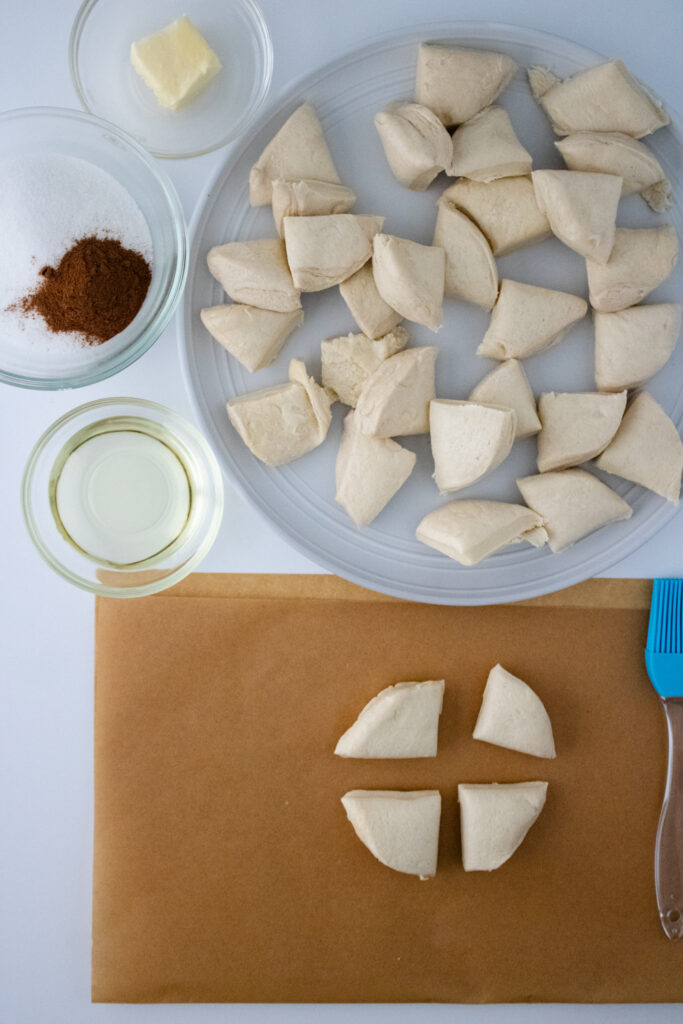 cut pieces of grands biscuit dough in a white plate along with four cut pieces placed on a brown paper towel with pastry brush aside, and vegetable oil, cinnamon power, butter in small glass cups