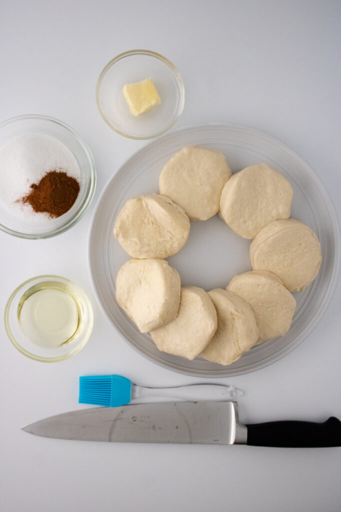 grands biscuit dough arranged in a glass plate along with butter, cinnamon powder, vegetable oil, pastry brush and knife
