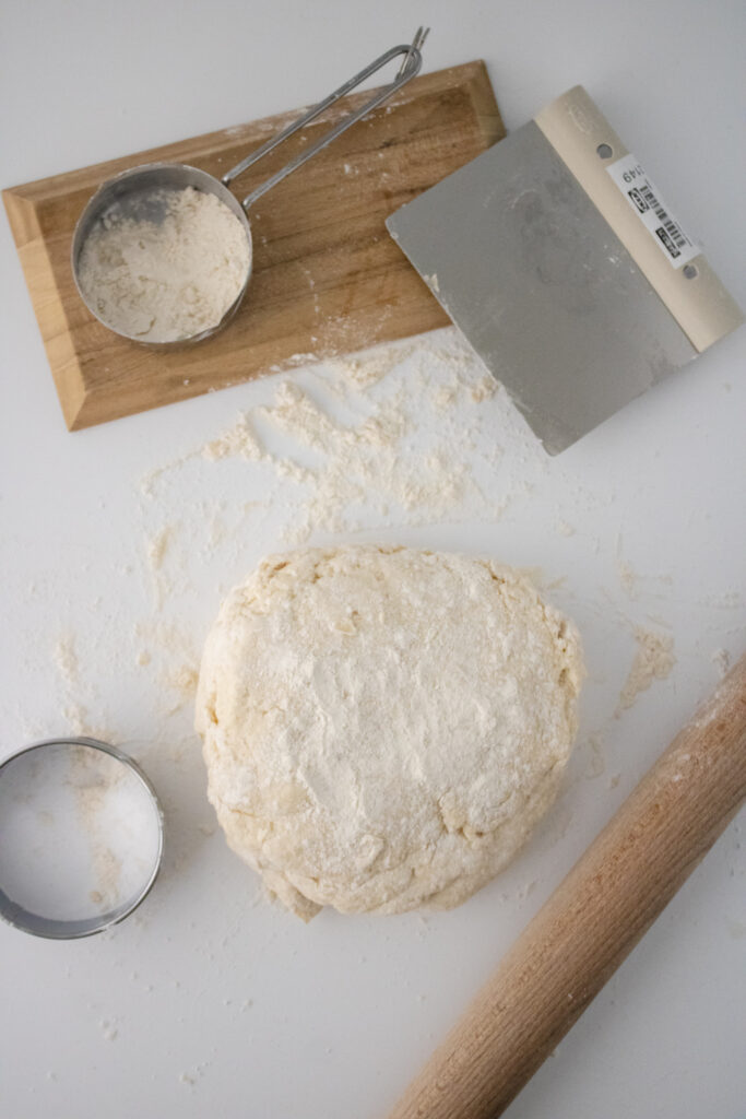wooden rolling pin, prepared dough, round cutter, bench scraper and a scoop of all-purpose flour
