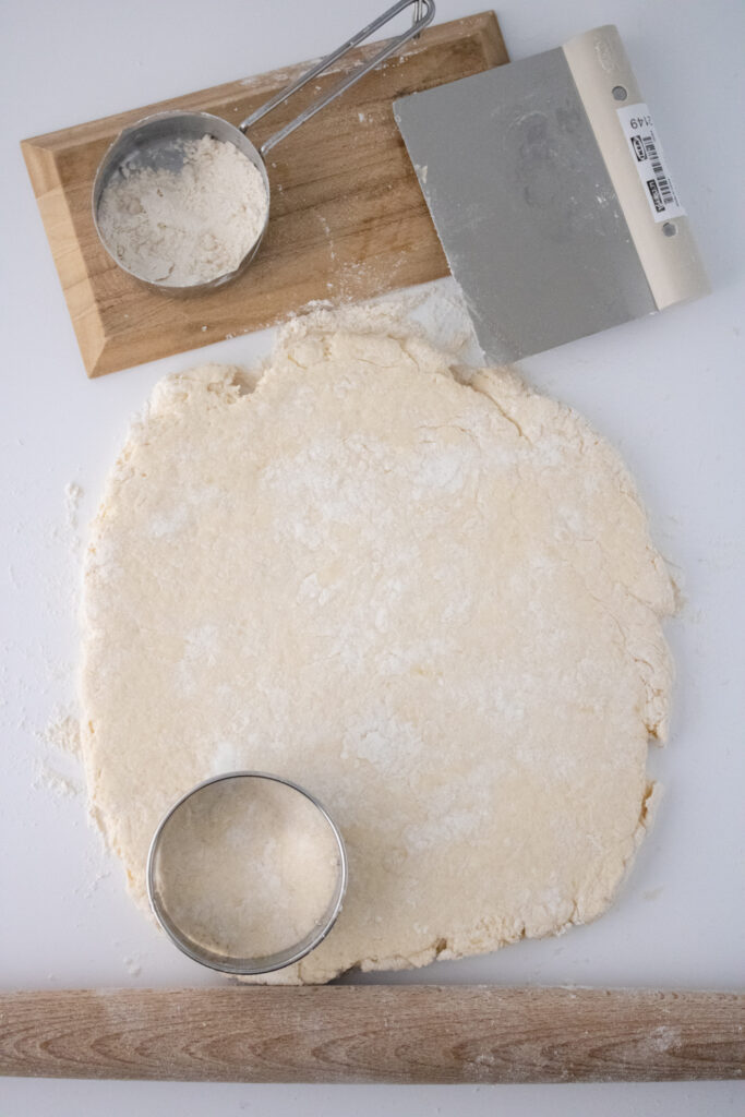 wooden rolling pin, round steel cutter placed on roll out dough along side bench scraper and a scoop of all-purpose flour