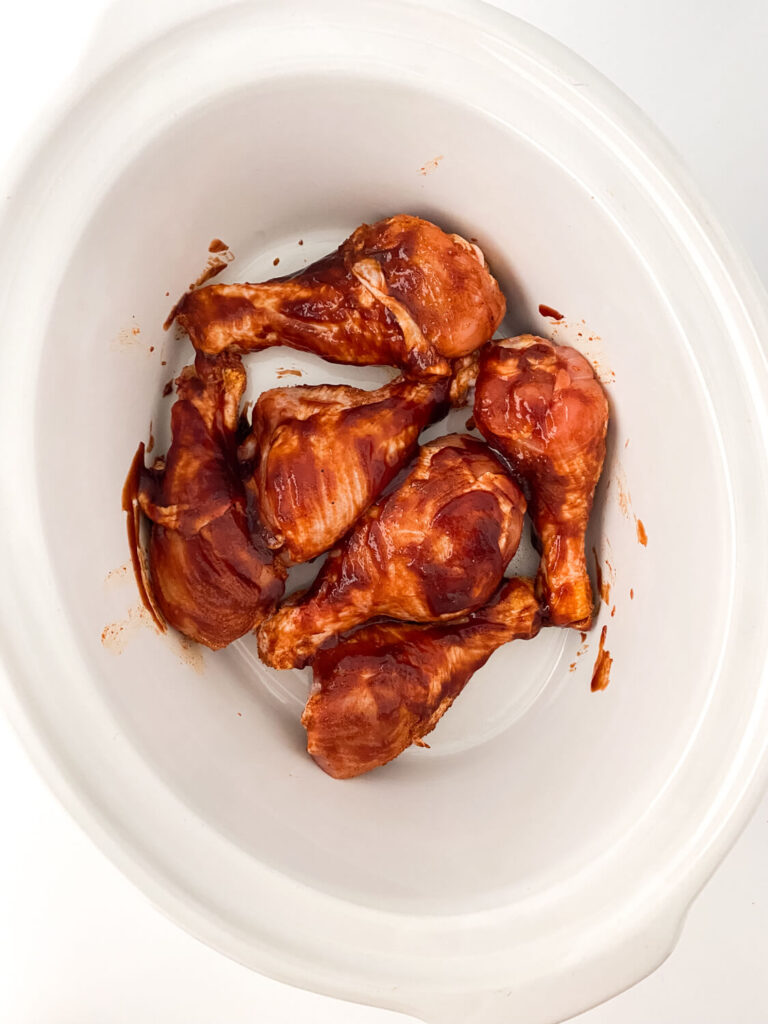 bbq sauce brushed evenly on seasoned chicken legs placed into the white crock pot