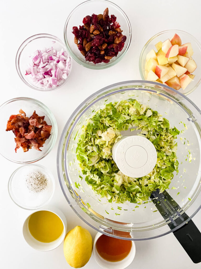 shredded Brussels sprouts in food processor along with chopped red onions, chopped fried bacon, candied pecans, dried cranberries, honey crisp apple in small glass bowls
