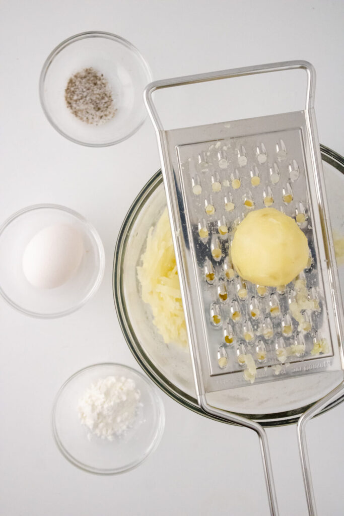 boiled peeled potato on grater on top of glass bowl along with pepper, egg, corn starch in small bowls