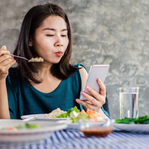 The girl is eating food while using her mobile phone, and she is wearing a blue dress
