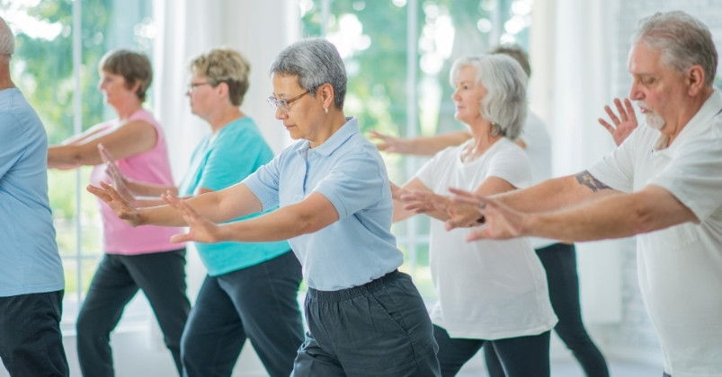 A group of older adults is practicing Tai Chi, moving slowly with their arms extended