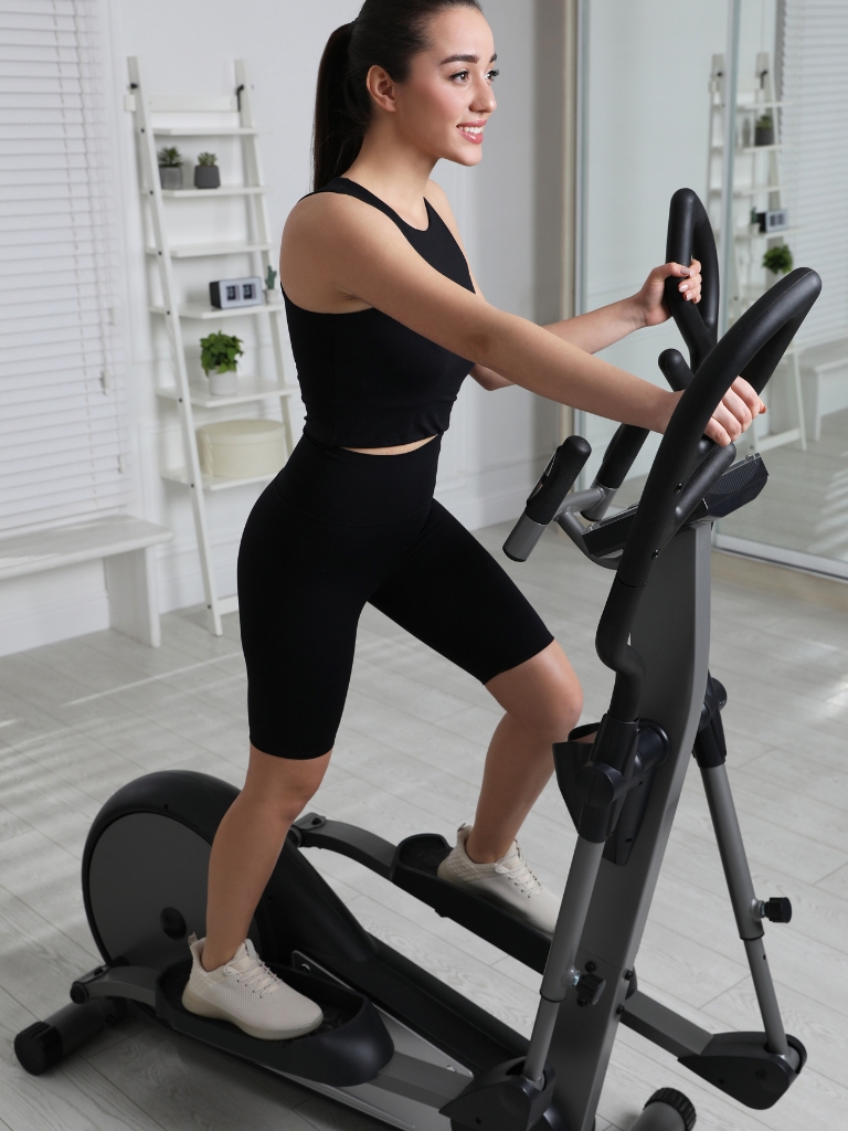 A woman exercises on an elliptical machine, wearing black athletic wear, in a minimalist room