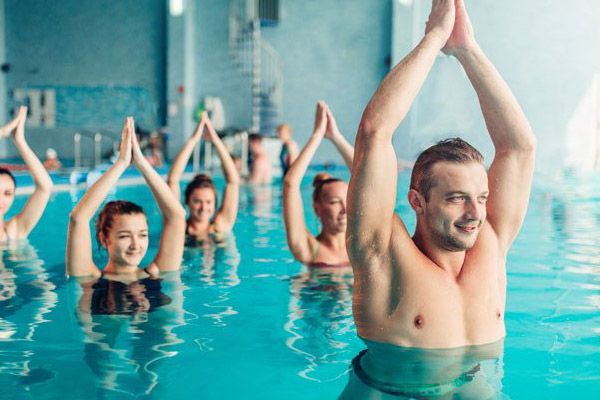 A group of men and women in a pool performs water aerobics, raising their hands overhead in a yoga-like pose