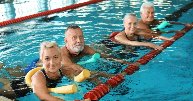 Four older adults engage in aqua fitness, using pool noodles for support as they float and move along the pool lane, smiling