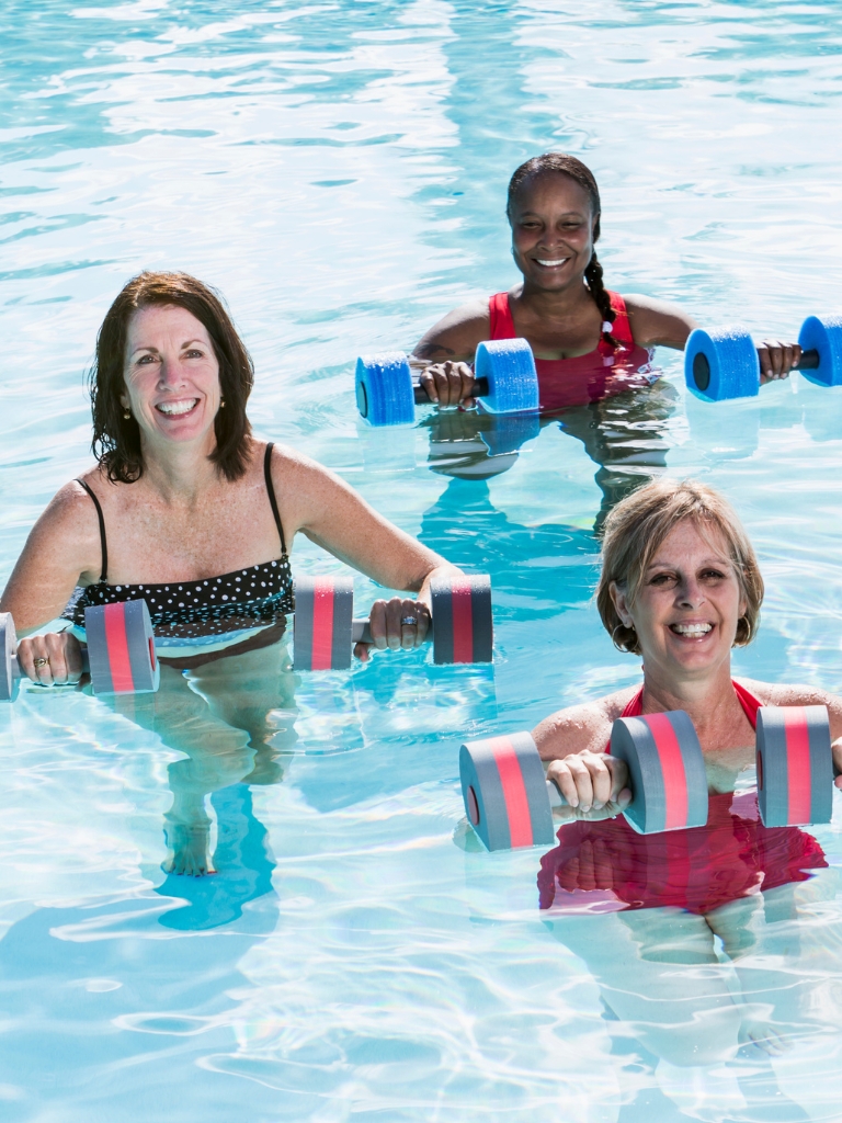 Three women are participating in a water aerobics class, standing in a swimming pool holding foam dumbbells