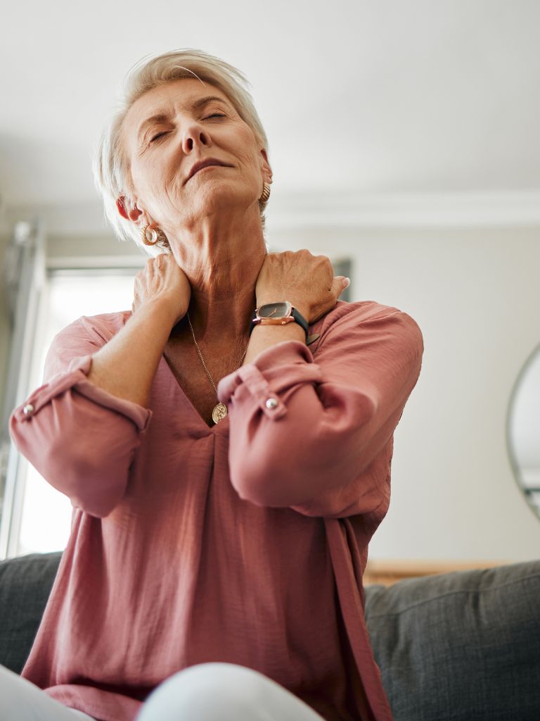 older woman experiencing neck pain. She is seated on a couch, holding her neck with both hands, massaging the area to relieve tension