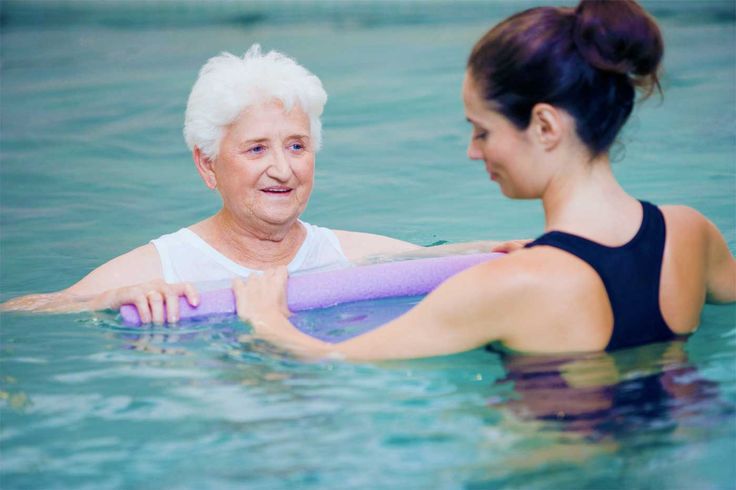 senior woman being assisted in an aquatic therapy session, holding a foam noodle for support while the instructor provides guidance